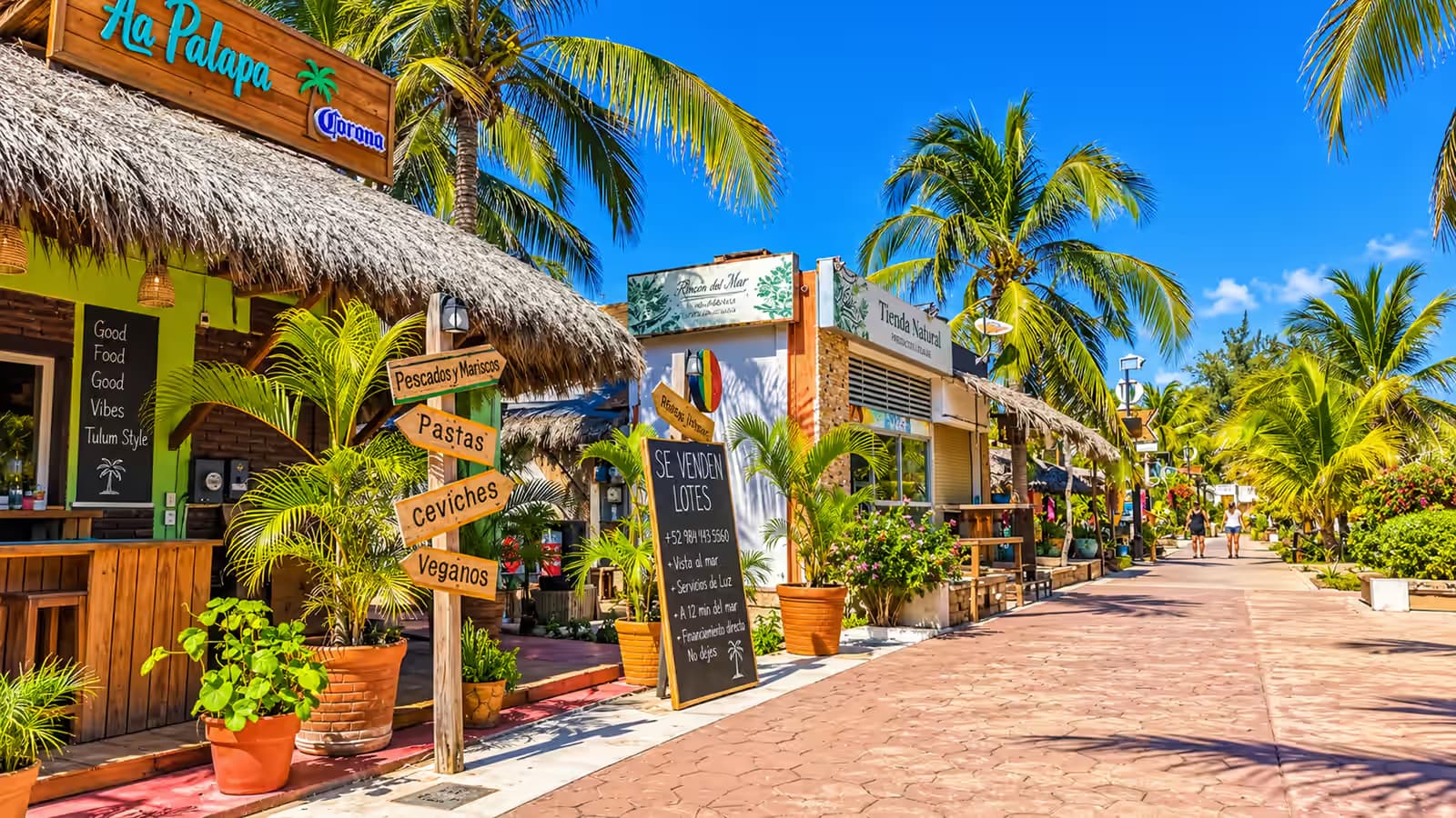 Calle peatonal con palapas, palmeras y letreros de restaurantes bajo un cielo despejado