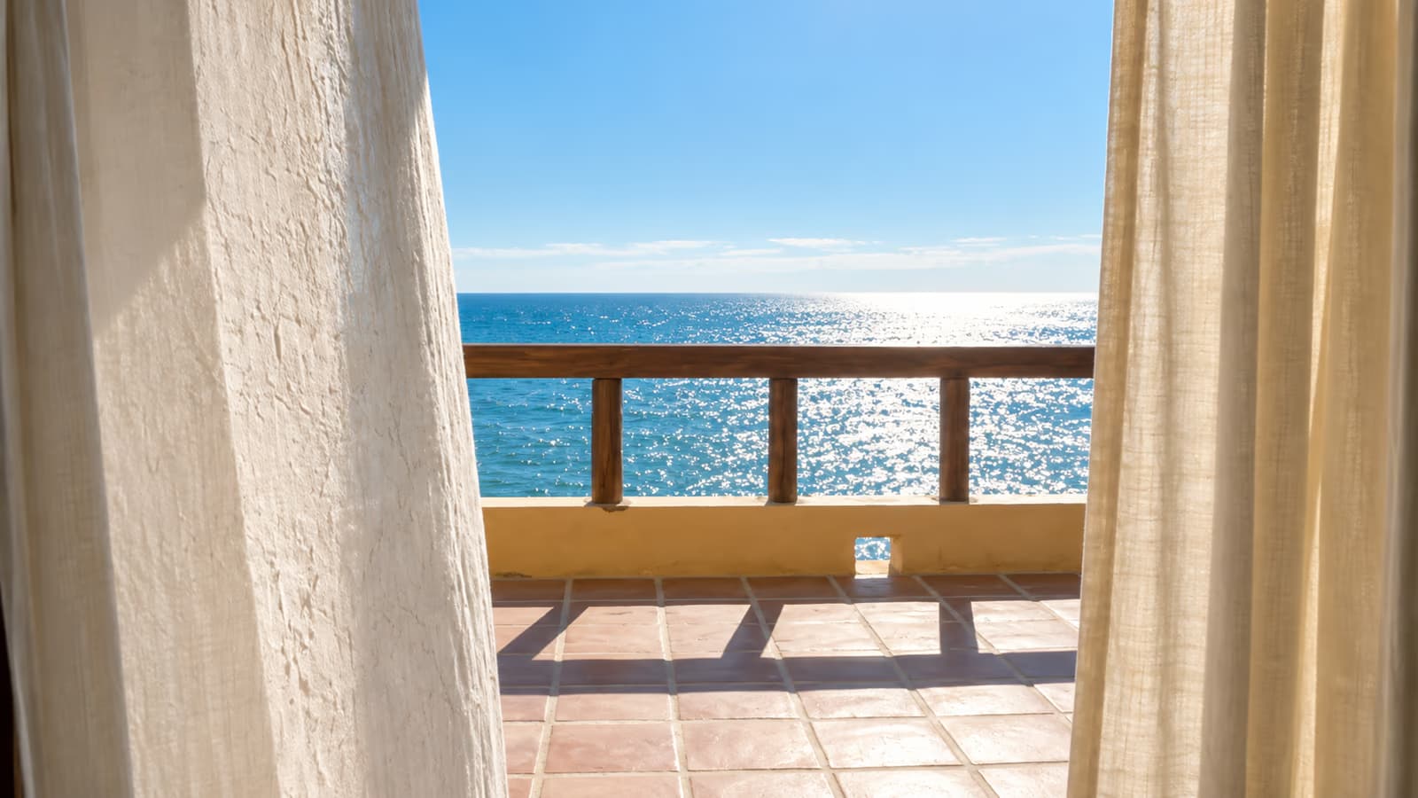 Terraza de piso de barro con cortinas de lino y barandal de madera frente al mar