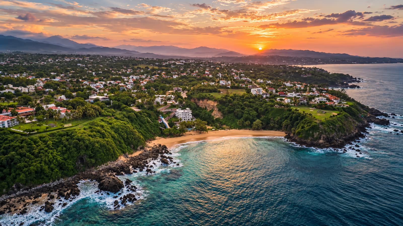 Aerial view of the coast at sunset, with houses among vegetation and the sun over the mountains